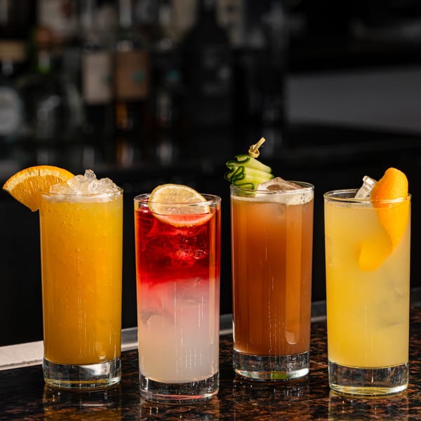 Four colorful cocktails with garnishes, including orange, lemon, and cucumber, on a bar.