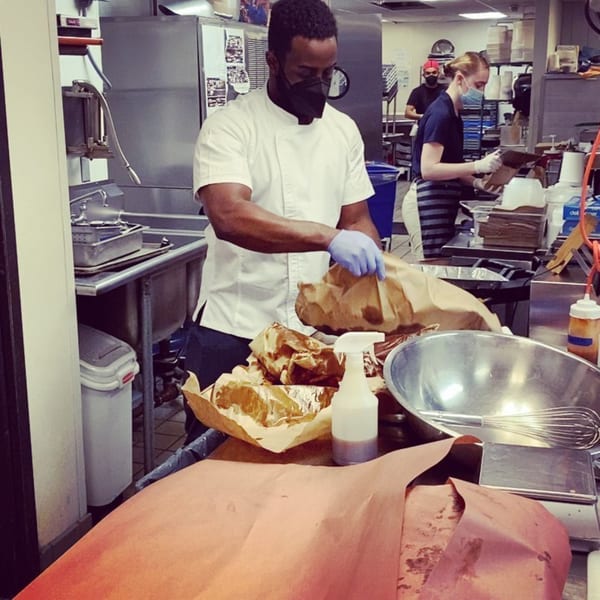 A chef in a face mask and gloves prepares food in a busy commercial kitchen at All Set Restaurant & Bar.