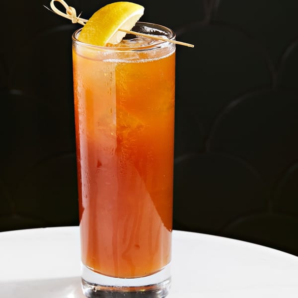Tall glass of orange cocktail with ice, garnished with a lemon wedge and a decorative pick.
