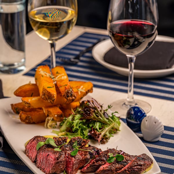 Sliced steak, golden fries, and a side salad on a white plate with wine glasses.