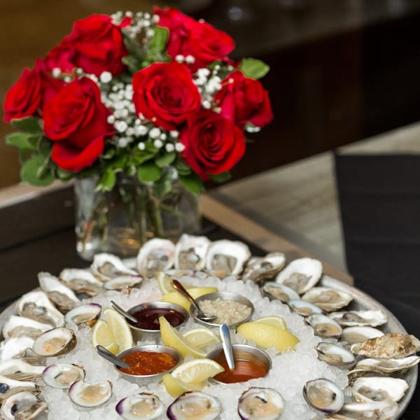 A platter of oysters and clams on ice with lemon wedges and sauces, next to a vase of red roses.