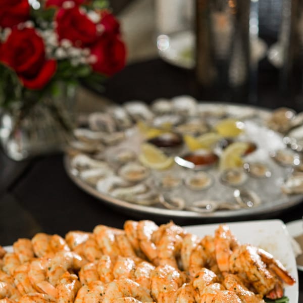 Plates of grilled shrimp and oysters on ice with lemon wedges, with a bouquet of red roses.