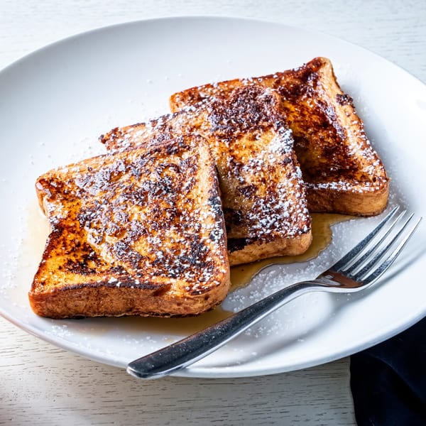 Three slices of French toast with powdered sugar and syrup on a white plate with a fork.