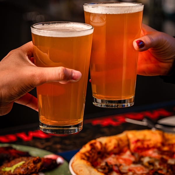 Two hands clinking glasses of amber beer, with a pizza and another dish blurred in the foreground.