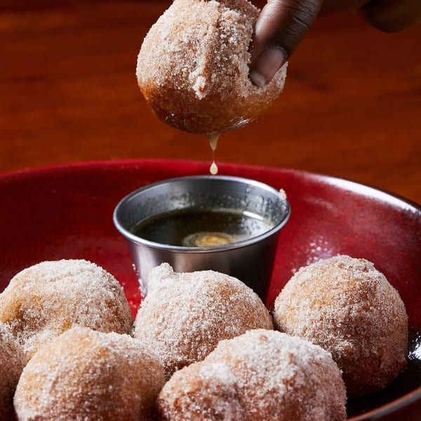 A hand dips a sugar-coated donut hole into a small metal cup of sauce on a red plate.