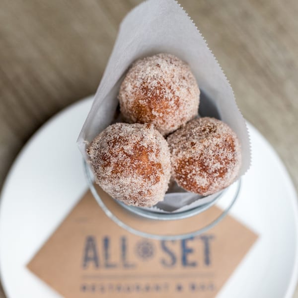 Three sugar-dusted donut holes in a metal bucket with a white paper liner, on a plate.