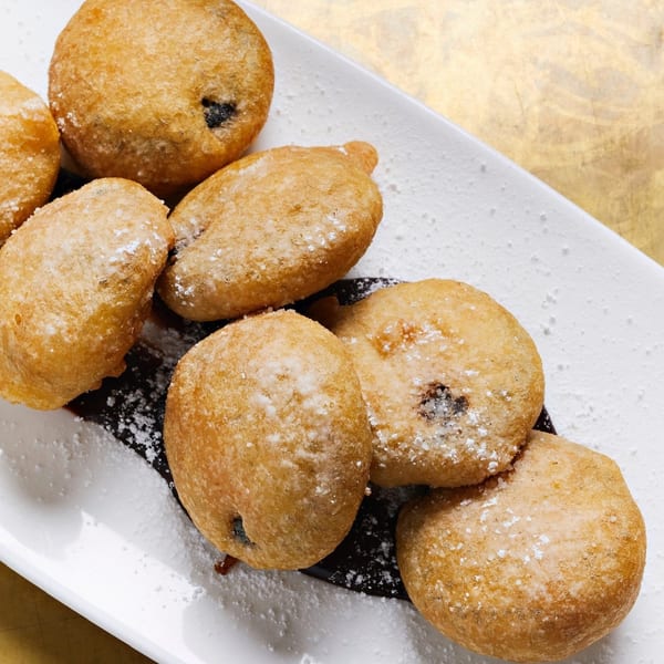 Golden-brown fried dough balls, dusted with powdered sugar, on a white plate with chocolate sauce.