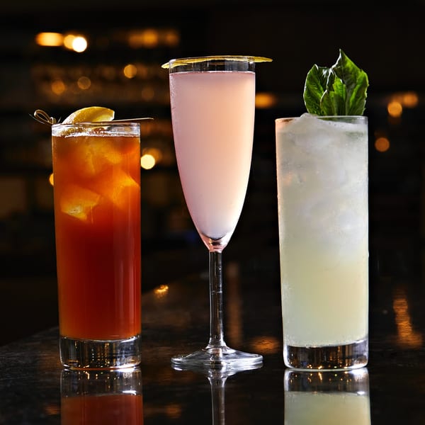 Three colorful cocktails, one red, one pink, and one light green, on a dark bar with bokeh lights.