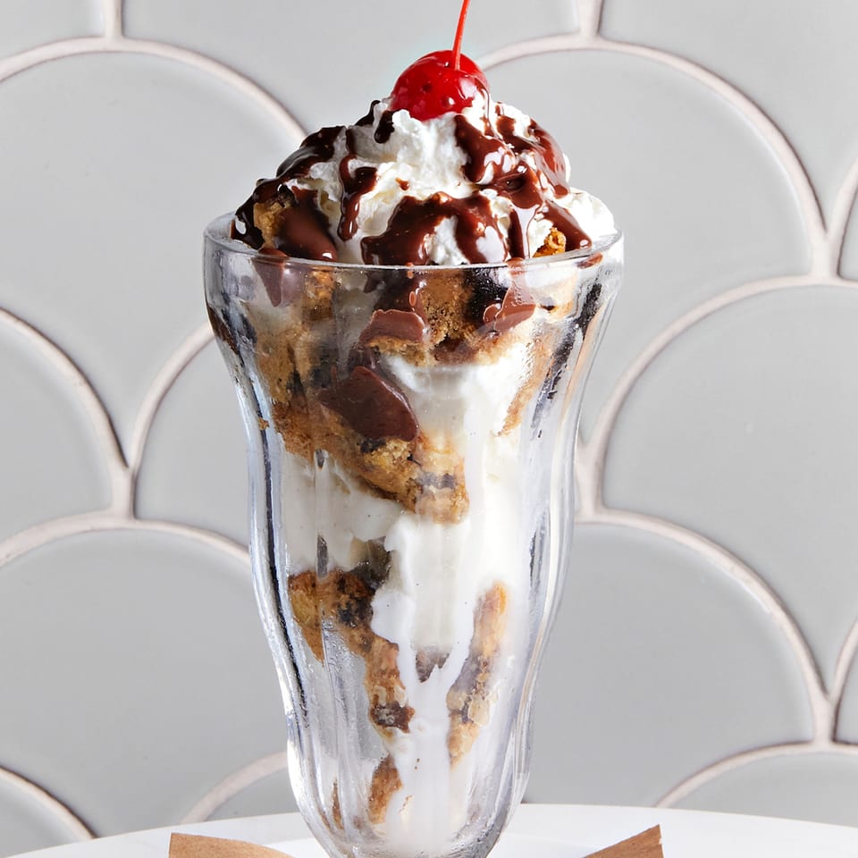 A tall glass of ice cream with cookie pieces, chocolate syrup, whipped cream, and a cherry.