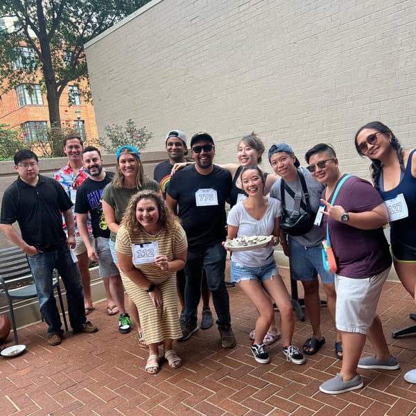 A group of smiling people, some holding oyster plates, pose outdoors on a brick patio.
