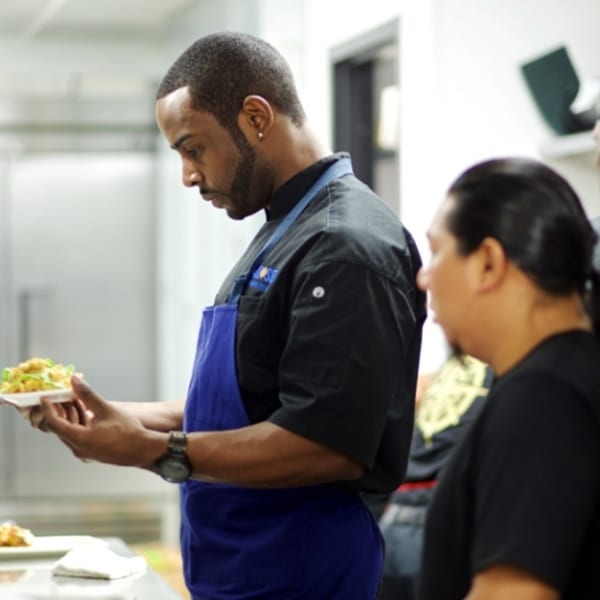 A chef in a black uniform and blue apron carefully inspects a plate of food.