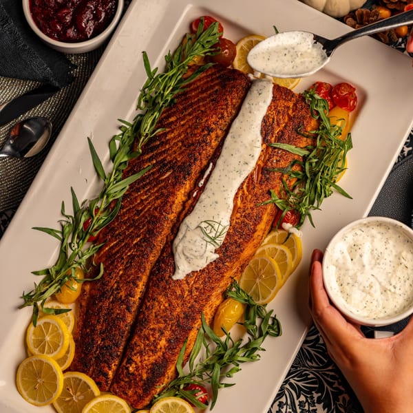 A large, seasoned salmon fillet on a white platter, garnished with lemon slices, herbs, and cherry tomatoes.
