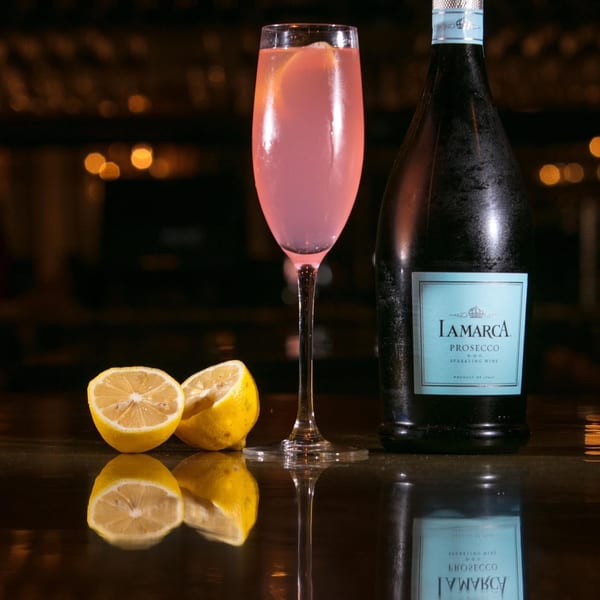 Pink cocktail in a flute glass, a bottle of La Marca Prosecco, and cut lemons on a bar.