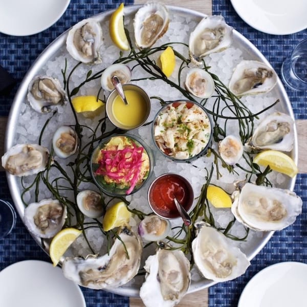 A seafood platter with oysters, clams, crab, and sauces on ice with lemon wedges.