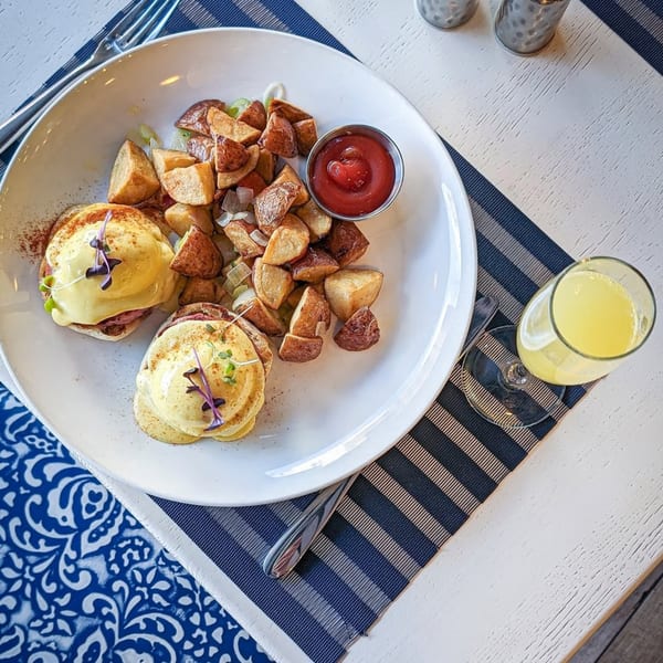 Two eggs Benedict with home fries, ketchup, and a mimosa on a striped placemat at All Set Restaurant & Bar.