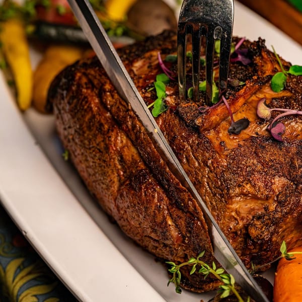 A large roasted meat dish, possibly prime rib, with a fork and knife, garnished with herbs.