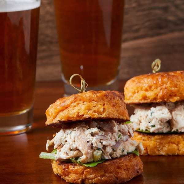 Two crab cake sliders on biscuits with lettuce, served with two glasses of beer.