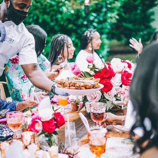 A server wearing a mask and gloves presents a platter of fried seafood to guests at an outdoor table.