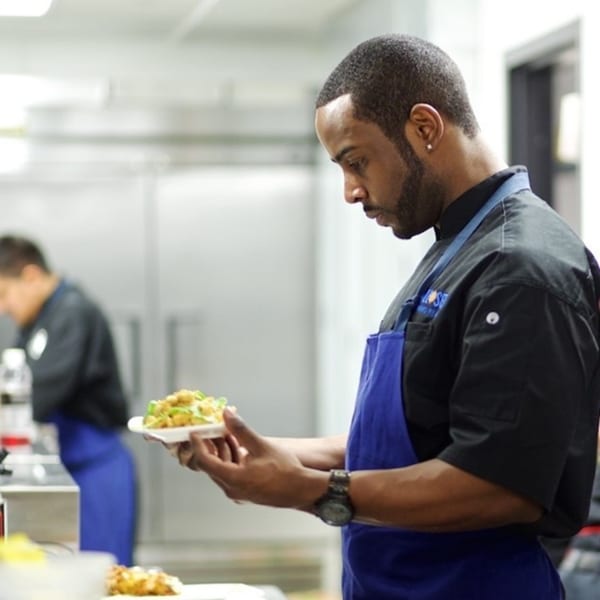 All Set chef in a black uniform and blue apron carefully garnishing a seafood dish.