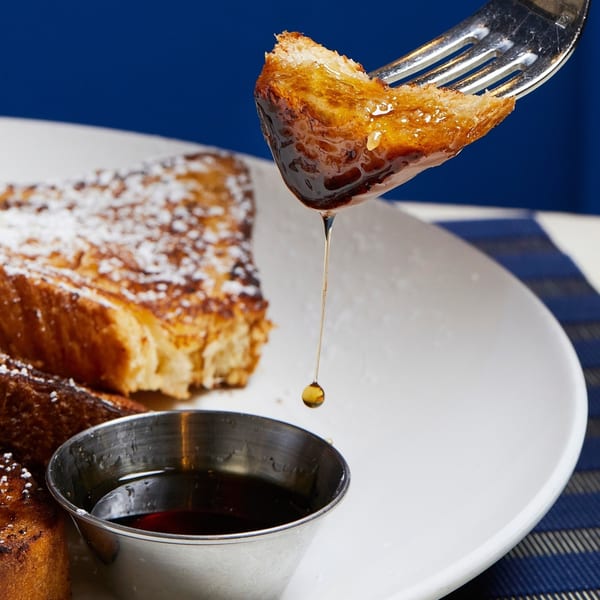 A fork lifts a piece of French toast, dripping syrup into a small metal cup on a white plate.