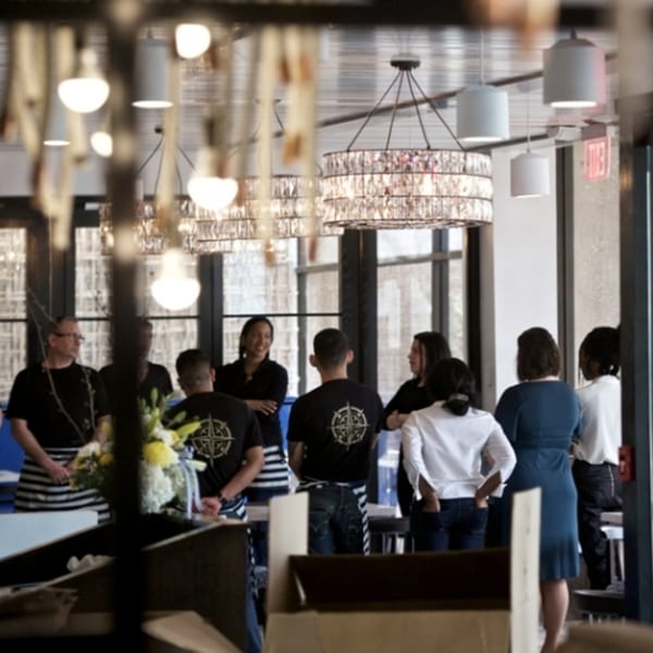 Restaurant staff, some in striped aprons and compass logo shirts, gather in a modern dining area.