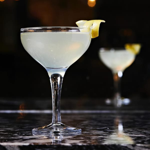 Two light-colored cocktails in coupe glasses with lemon twists on a dark, reflective bar.