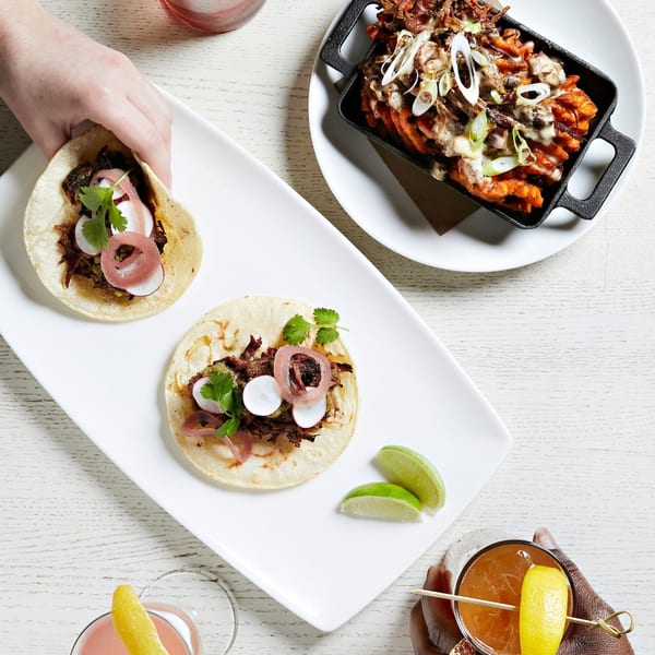 Overhead view of two hands, one holding a taco, with another taco and a skillet of fries.
