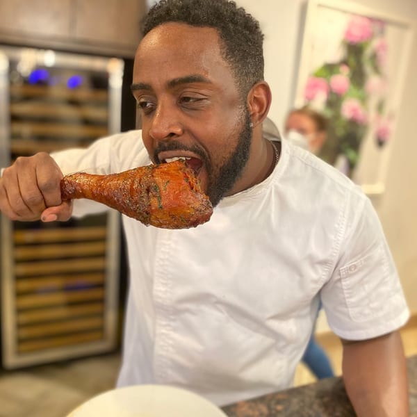 A man in a chef's coat enthusiastically bites into a large, glazed chicken drumstick.