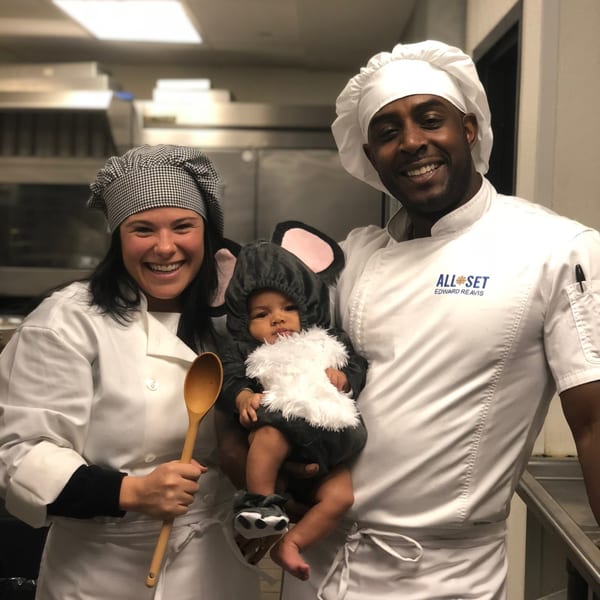 Two All Set chefs, a man and a woman, smiling and holding a baby in a mouse costume.