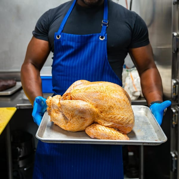 Chef in blue apron and gloves holding a raw, seasoned turkey on a metal tray.