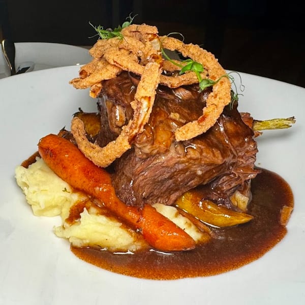 Braised short ribs with mashed potatoes, roasted carrots, and crispy fried onions on a white plate.