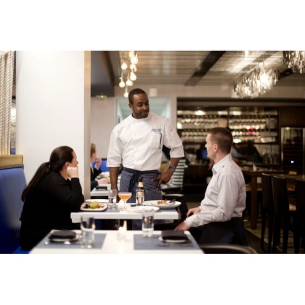Chef in white coat and striped apron talking to two customers at a table in All Set Restaurant.