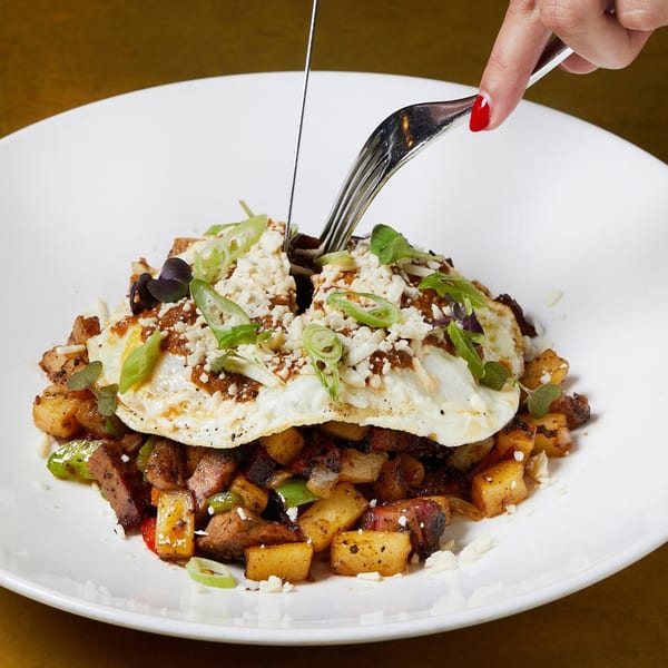 A hand cuts into a dish of fried eggs, potatoes, and meat, garnished with cheese and scallions.