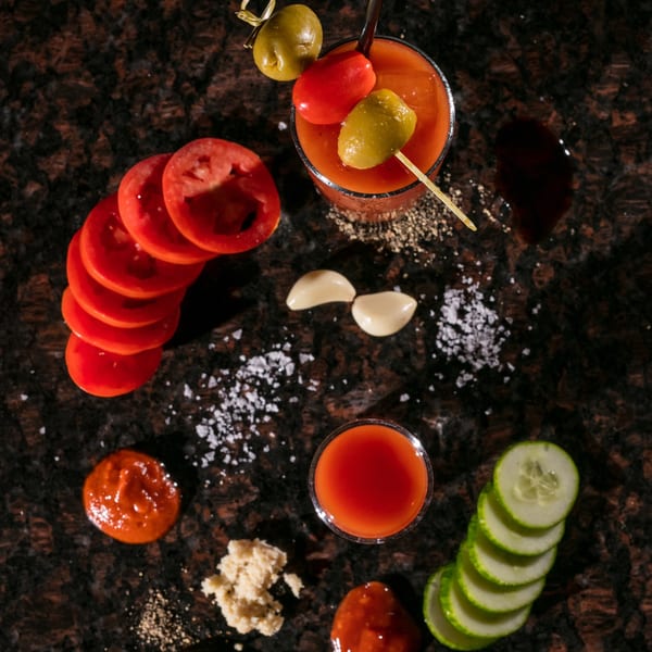 Overhead view of a Bloody Mary cocktail with garnishes, spices, and sliced vegetables on a dark surface.