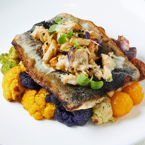 Pan-seared fish topped with crab meat, microgreens, and colorful cauliflower on a white plate.