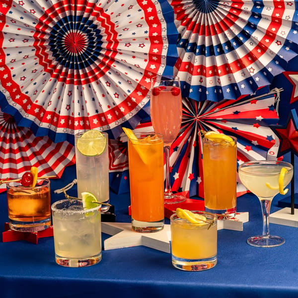 Seven colorful cocktails on a blue table with red, white, and blue star-patterned decorations.