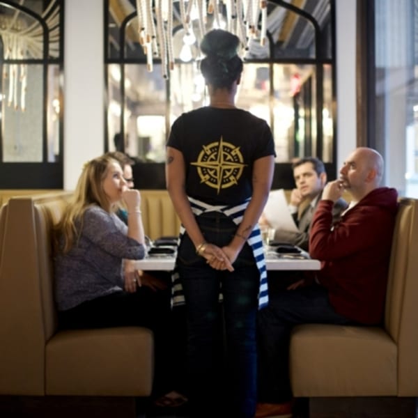A server with a compass logo shirt stands at a table with three seated customers in a restaurant.